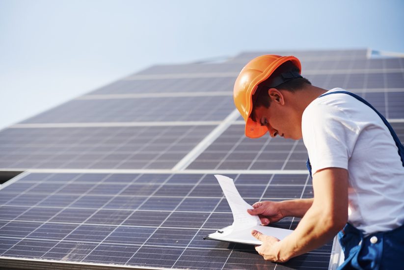 With documents. Male worker in blue uniform outdoors with solar batteries at sunny day